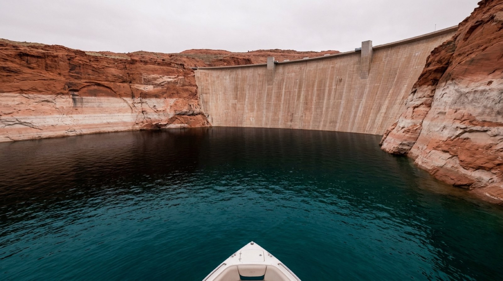 The curved concrete face of Glen Canyon Dam holding back deep blue-green water in a narrow red sandstone canyon, pale mineral bathtub rings high on both canyon walls marking former higher waterlines, seen low from a small boat under flat overcast light.