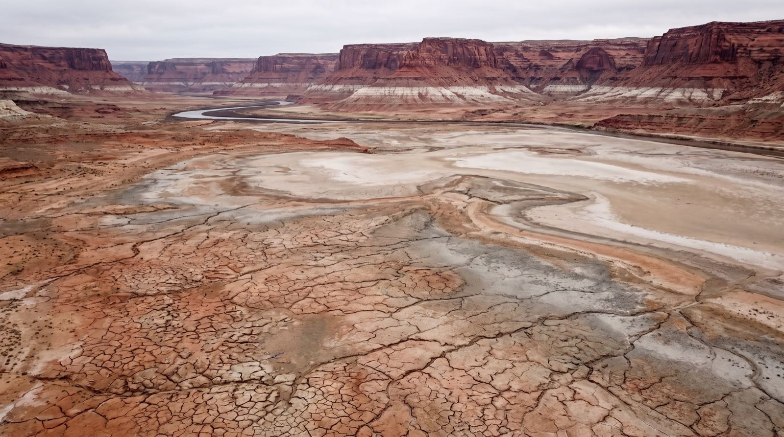A wide expanse of cracked, sun-baked former lakebed and pale exposed sediment flats stretching toward a distant thin ribbon of Lake Powell water beneath red sandstone cliffs