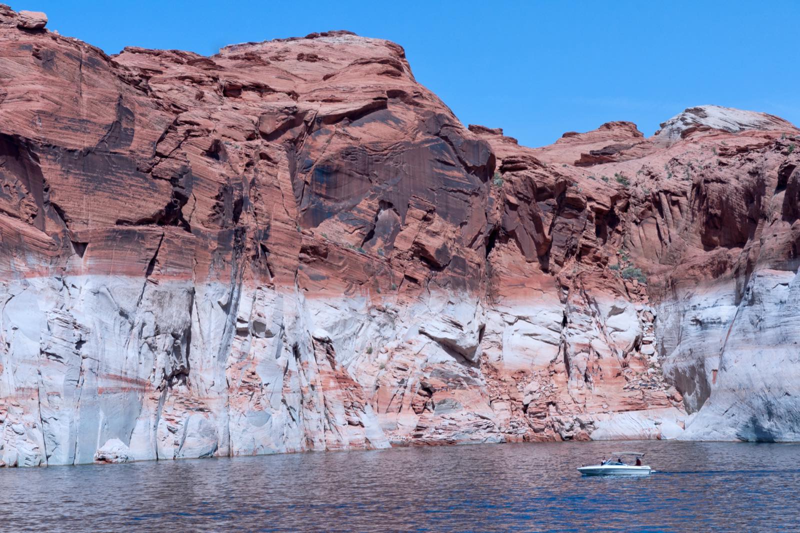 Red sandstone cliffs at Lake Powell above a wide band of bleached white rock marking the old high-water line, with a small boat on the water below