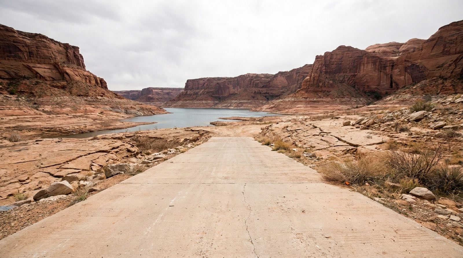 A pale concrete Lake Powell boat launch ramp ending on cracked, dry lakebed, the reservoir's water receded far below and beyond the end of the ramp, red sandstone cliffs behind under an overcast sky