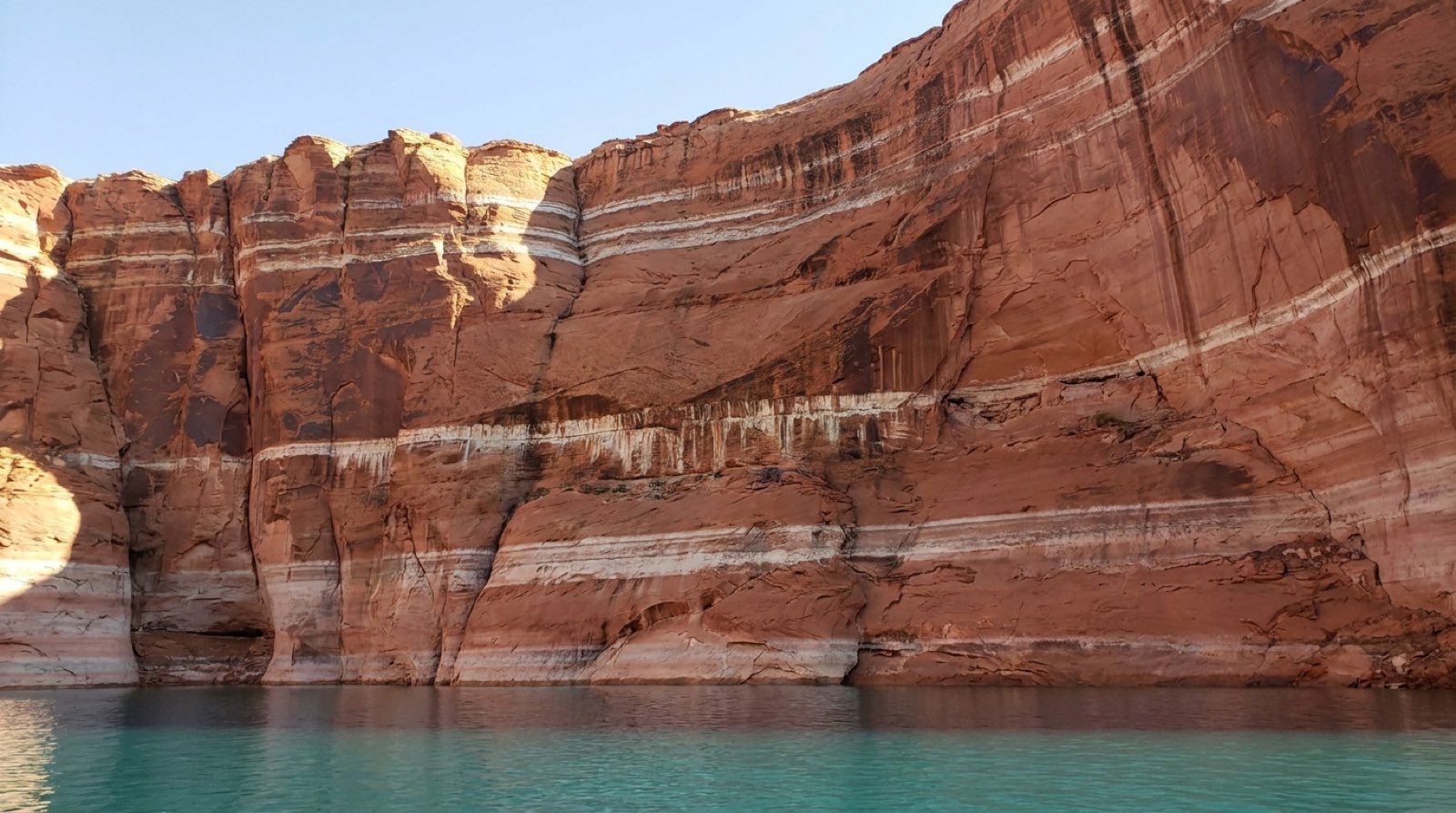 Layered pale mineral bands on Lake Powell's red sandstone canyon walls, each ring marking a former water line from a different year, with the current reservoir far below the highest band.