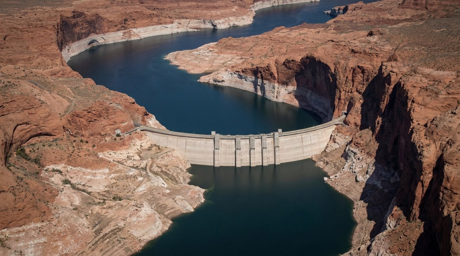 Glen Canyon Dam holding back a lowered Lake Powell, with a wide pale mineral bathtub ring on the red sandstone marking the 3,700-foot full-pool line far above the current water surface.