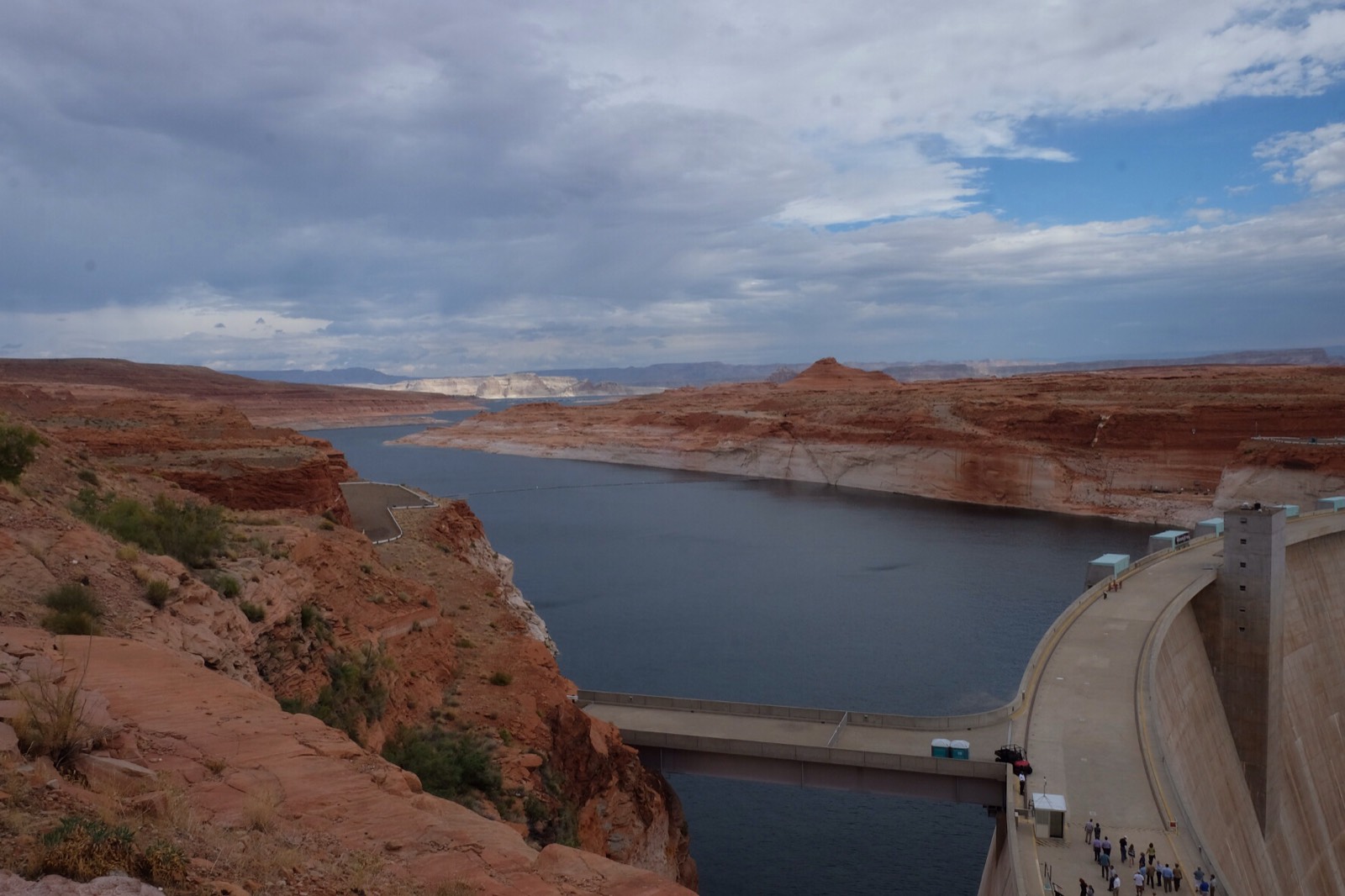 Glen Canyon Dam and the Glen Canyon Dam Bridge at the head of Lake Powell, the red sandstone canyon walls marked by a tall pale mineral bathtub ring far above the low reservoir surface under an overcast sky