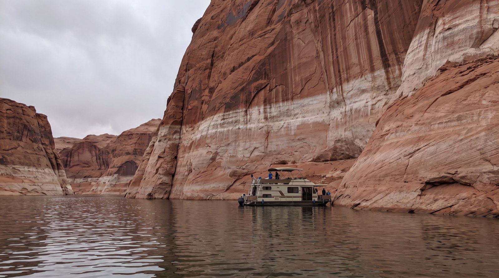 The pale mineral bathtub ring on Lake Powell's red sandstone canyon walls towering far above the current waterline, a small houseboat at the base for scale