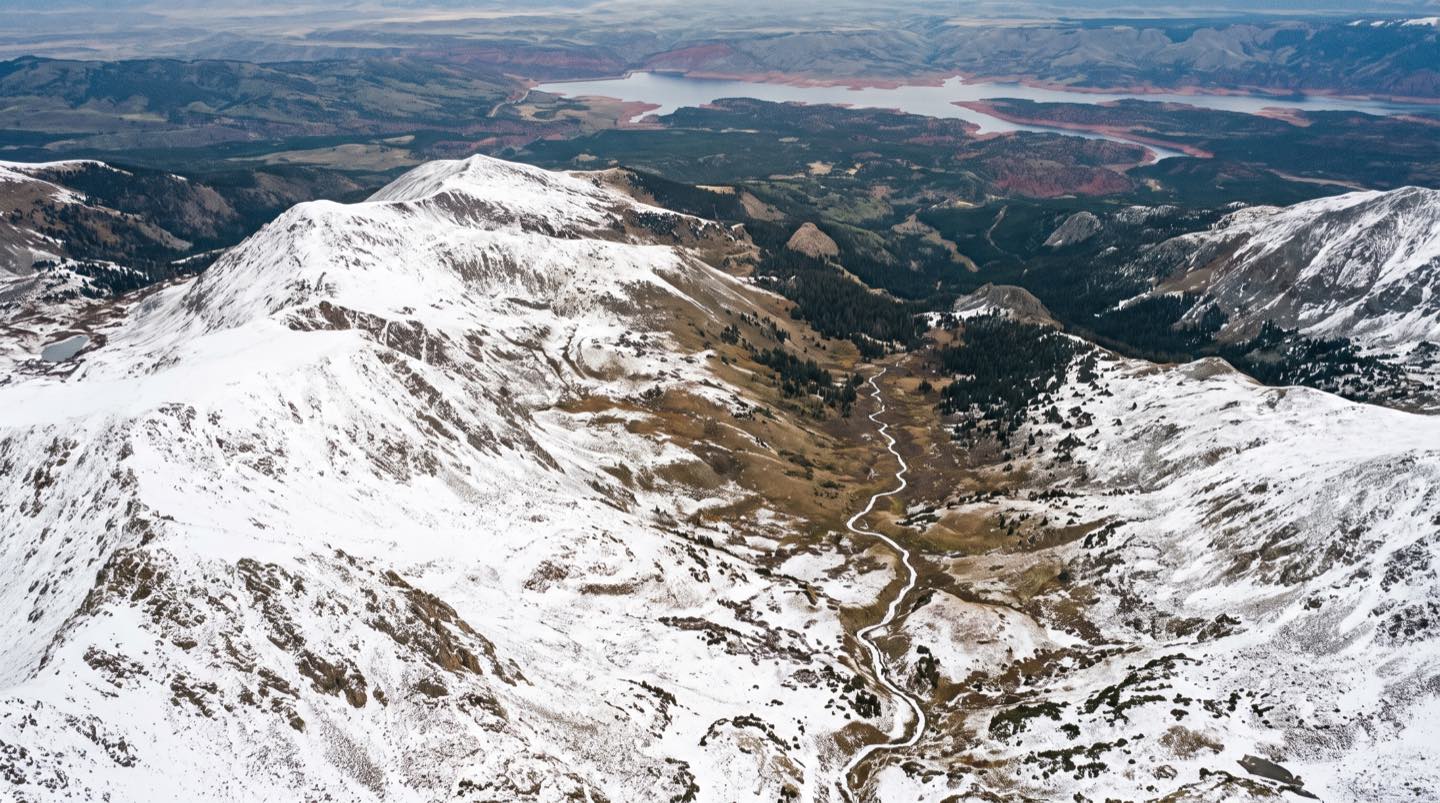 Snow-covered Rocky Mountain peaks feeding a thin meltwater stream that winds down toward a distant red-rock reservoir, seen from a high aerial perspective